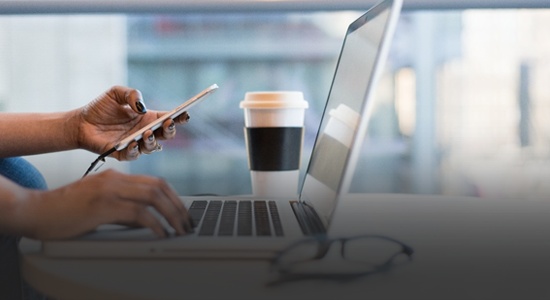 Someone using a laptop, on a desk - whilst holding a phone 