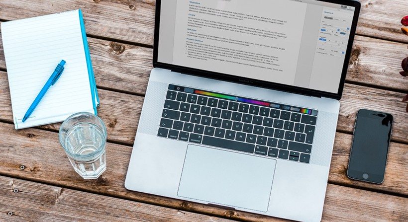 Laptop on a wooden desk, next to a phone and notepad