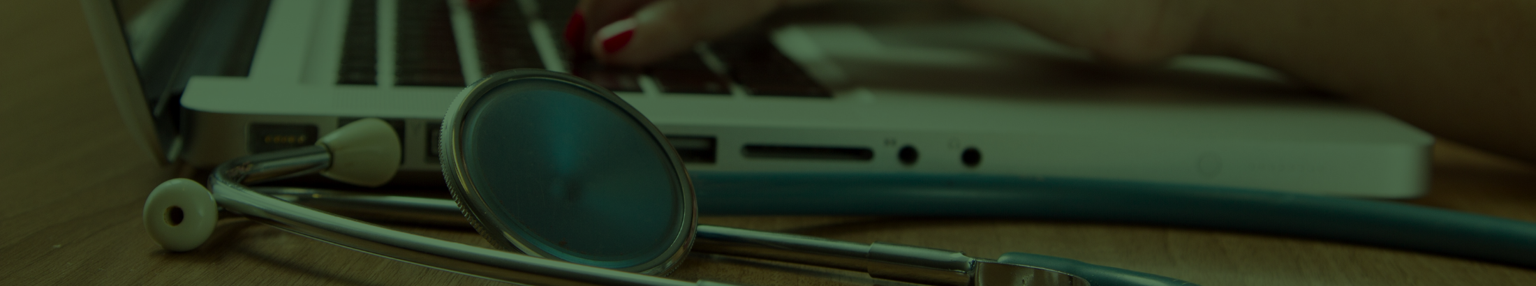 Hands typing on a laptop with a stethoscope by its side on the desk.