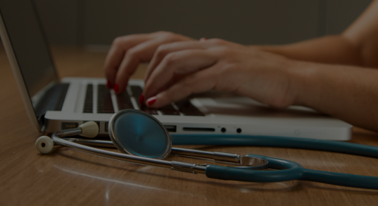 Hands typing on a laptop with a stethoscope by its side on the desk.