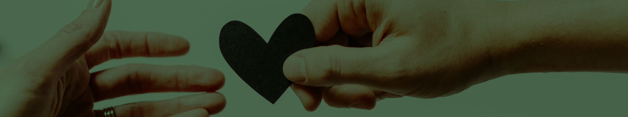 A photograph of a hand reaching across the frame, handing a paper cut-out of a heart to another hand.